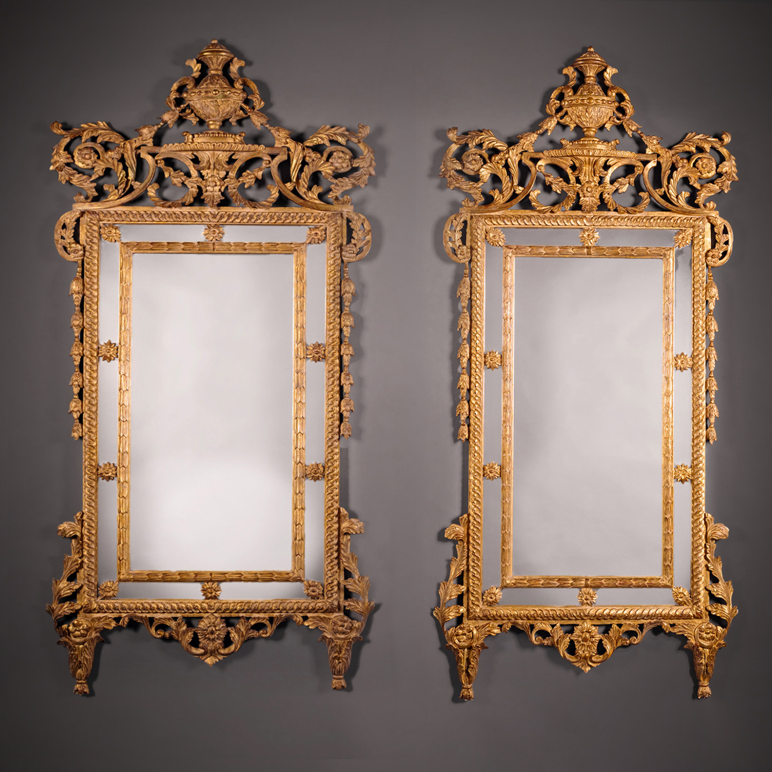 Pair of Italian neoclassical giltwood pier mirrors with rectangular plates, husk-carved slips, rosettes, scrolling foliage cresting, and urn finials, early 20th century.