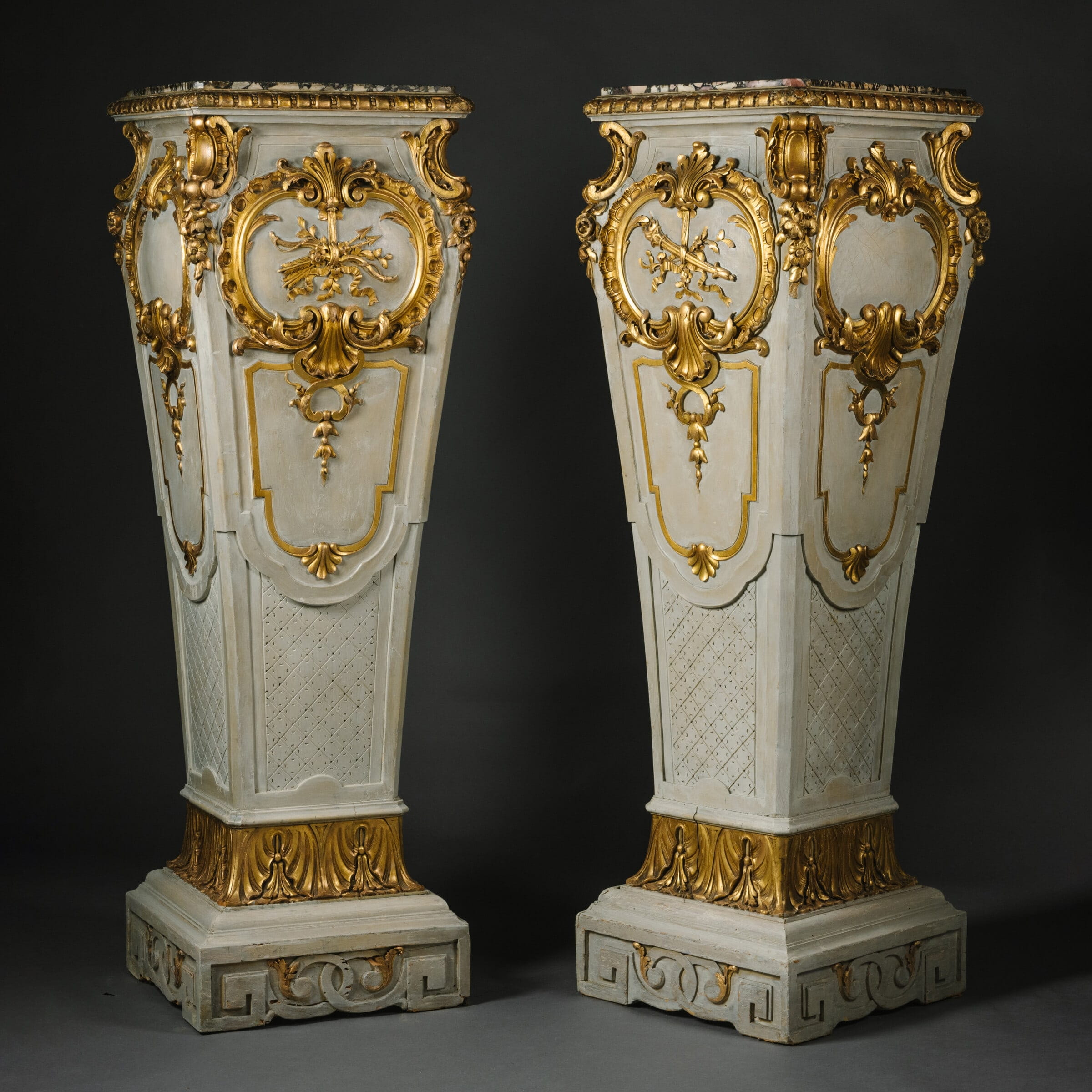 Pair of large parcel-gilt and grey painted pedestals, France circa 1900, featuring Escalette Alpha marble tops, carved scrollwork, acanthus cartouches, and Greek-key patterned bases.