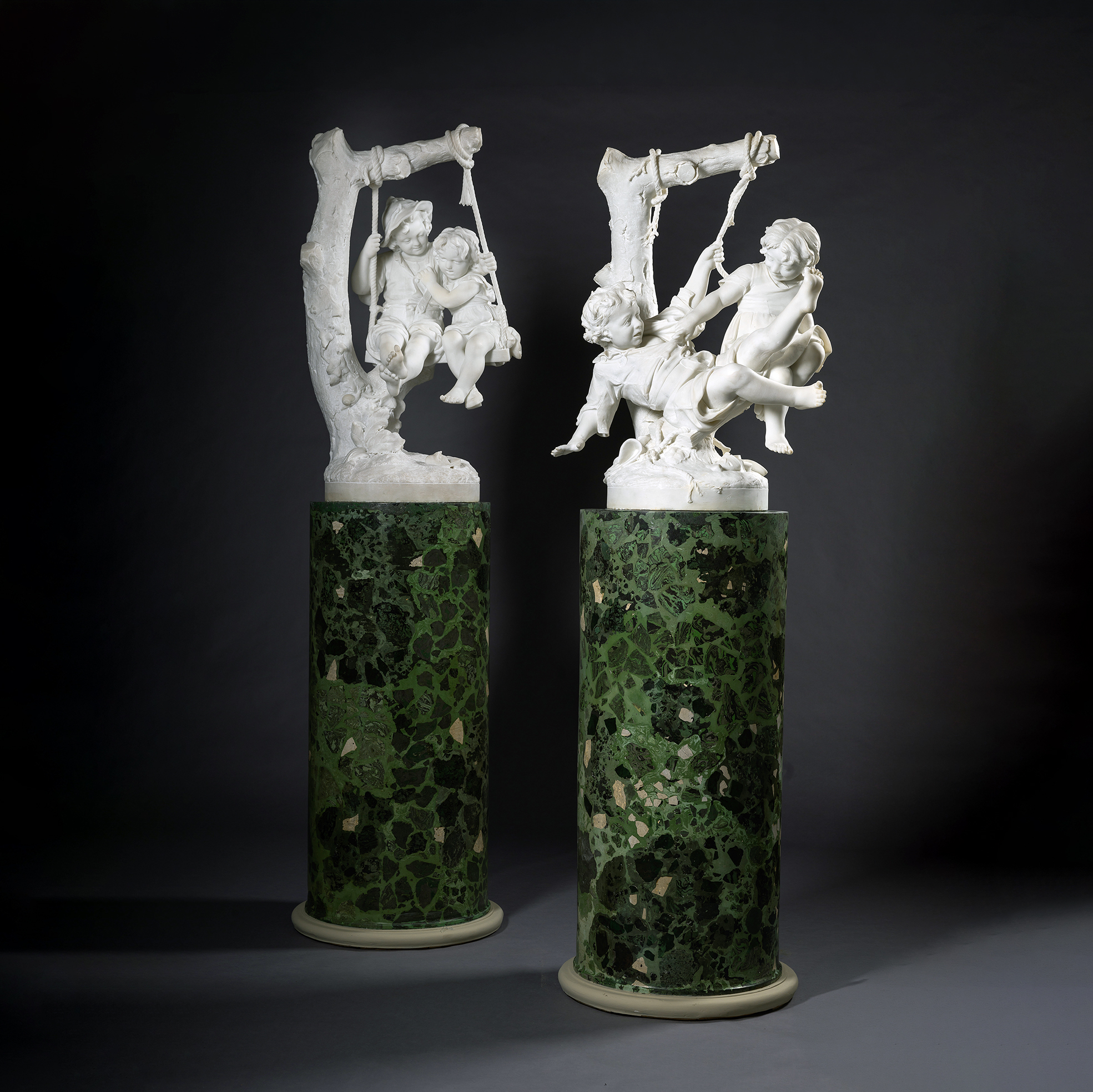 Pair of Carrara marble figural groups of children on swings by Alessandro Lazzerini, Italy, circa 1900, displayed on green scagliola pedestals.