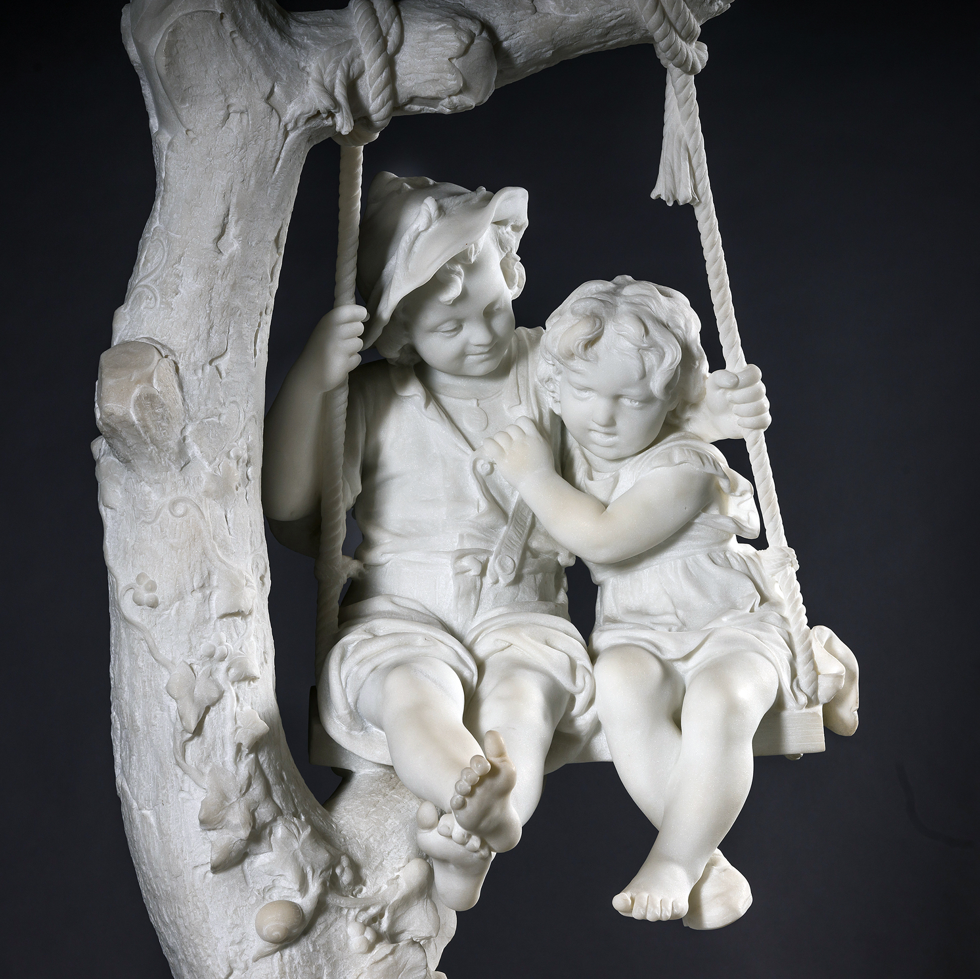 Carrara marble sculpture 'Two Children on a Swing' by Alessandro Lazzerini depicting a boy and girl seated on a swing beneath an ivy-clad tree.