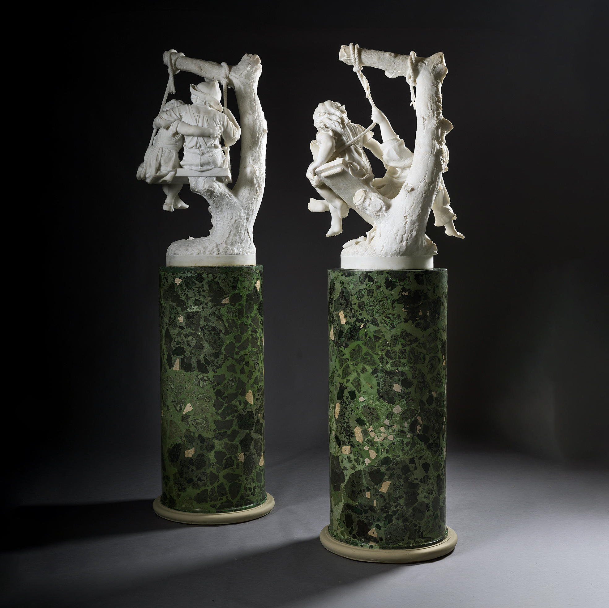 the back view of two marble figural groups of children on swings by Alessandro Lazzerini, Italy, circa 1900, displayed on green scagliola pedestals.