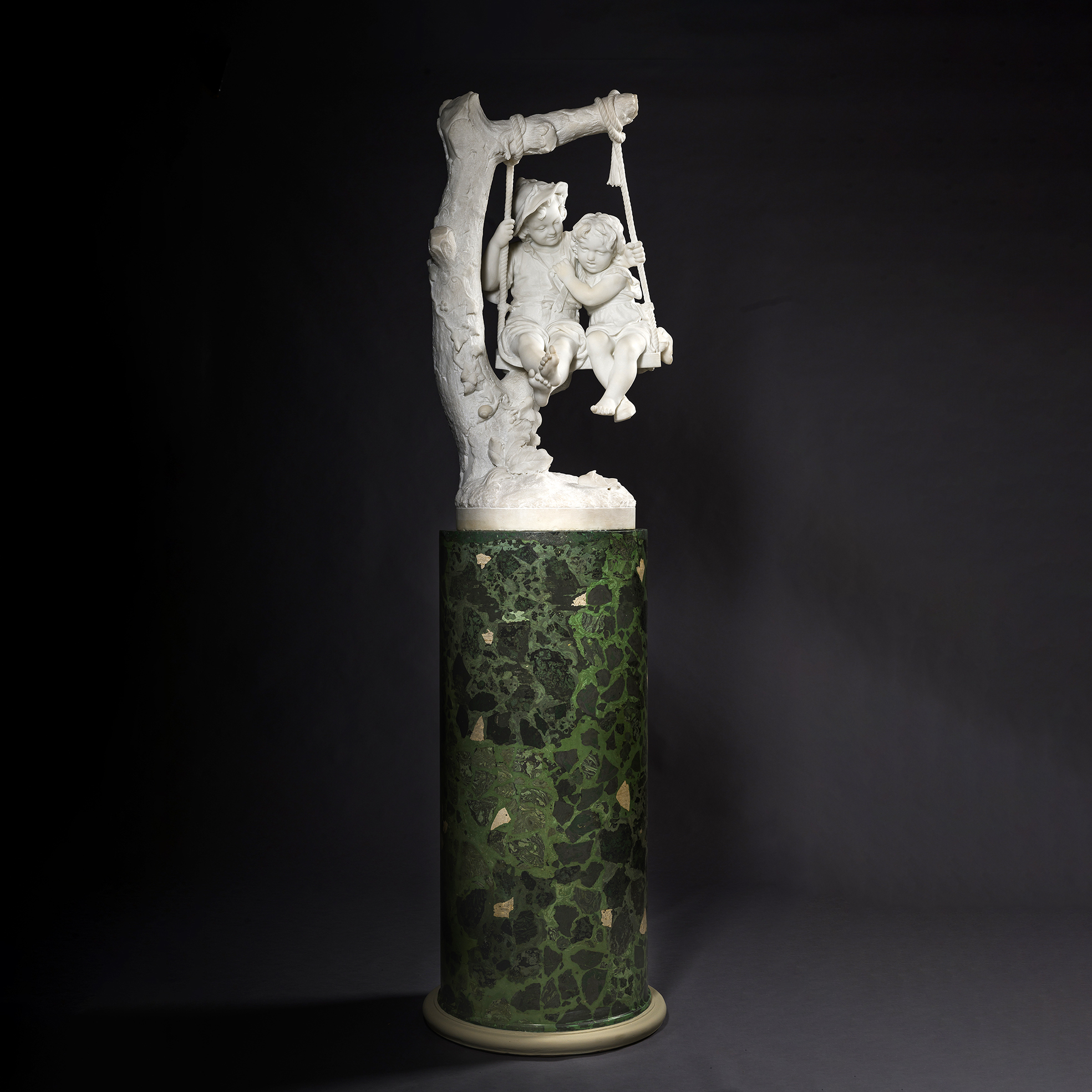 Carrara marble sculpture 'Two Children on a Swing' by Alessandro Lazzerini depicting a boy and girl seated on a swing beneath an ivy-clad tree.